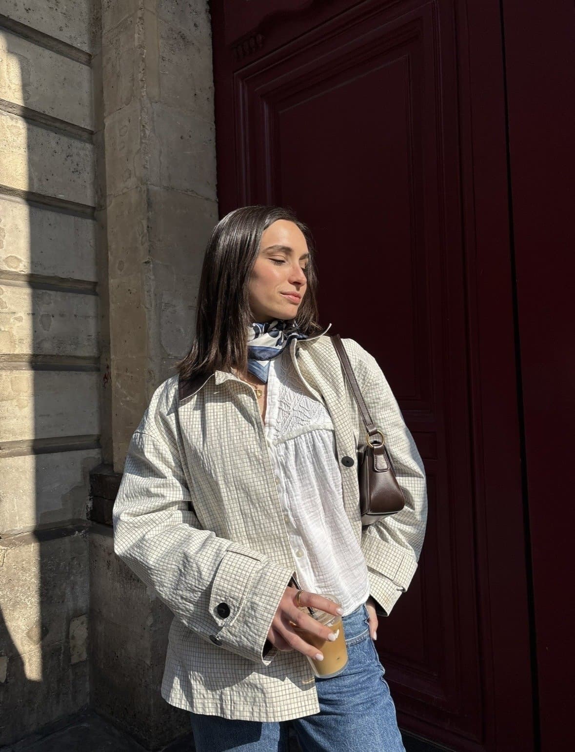 Oversized light checked jacket layered over a white textured blouse, blue jeans, a neck scarf, and a dark brown shoulder bag.