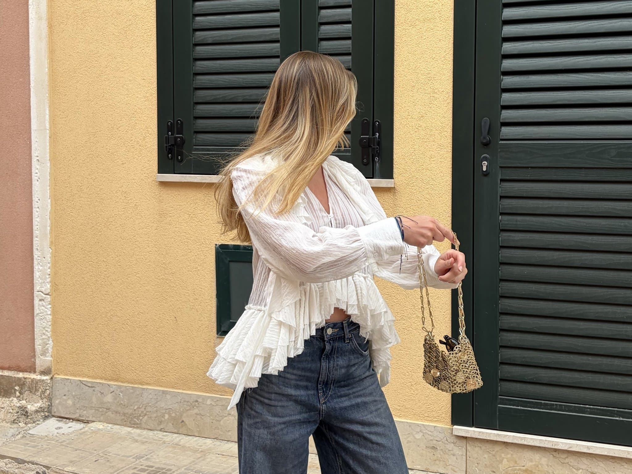 A white ruffled sheer blouse is styled with dark blue jeans, bracelets, and a small gold chain handbag.