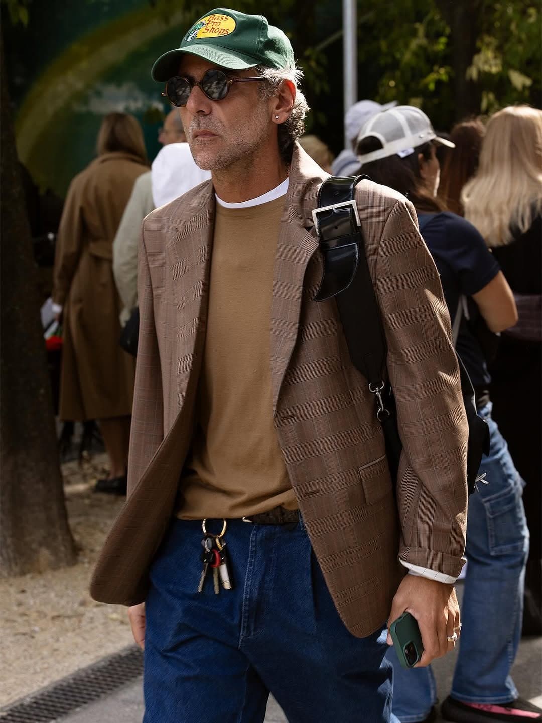 A brown plaid blazer is worn over a tan knit top with blue jeans, a green logo cap, round sunglasses, and a black shoulder bag.