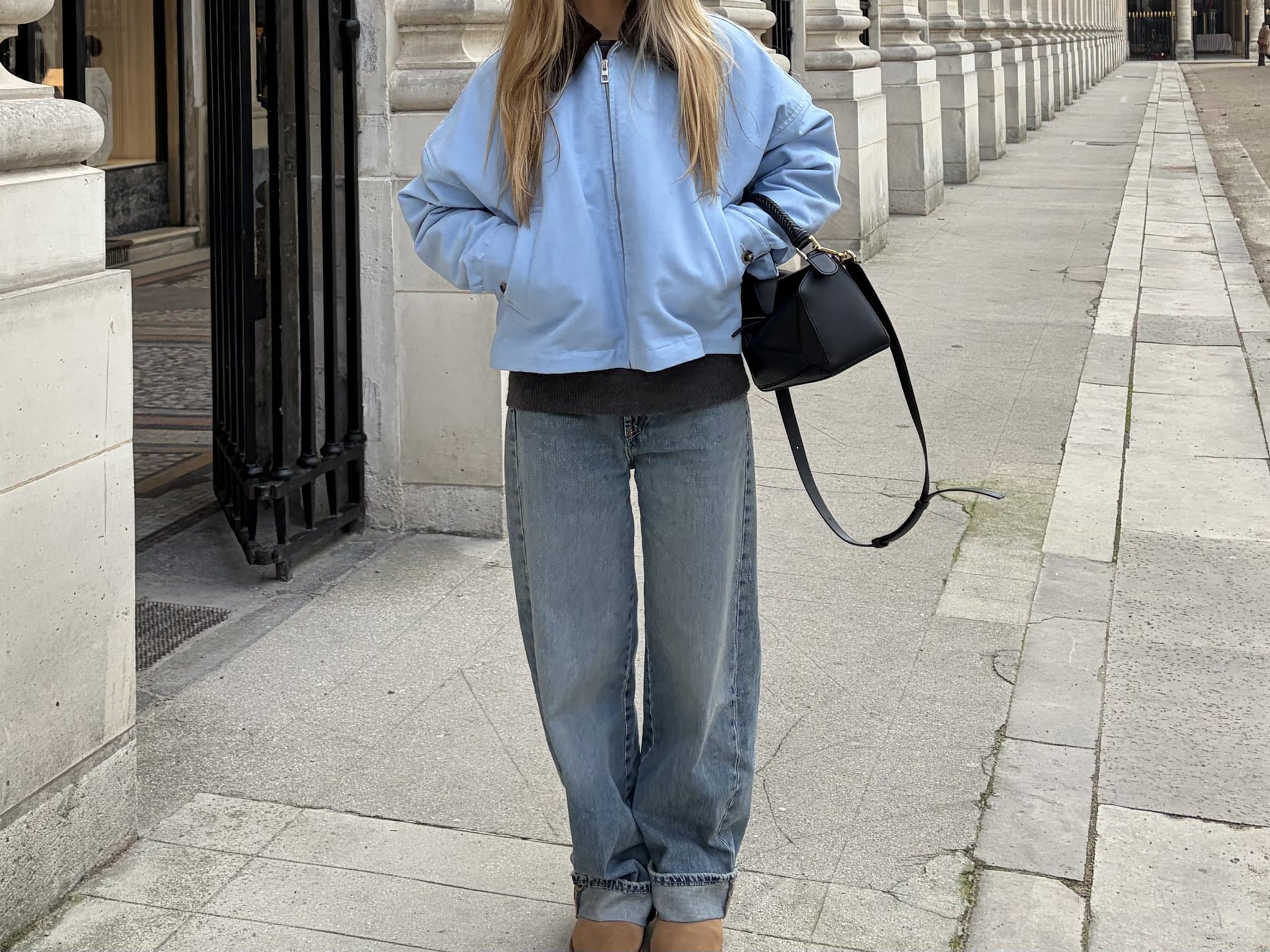 Oversized pale blue zip jacket layered over a dark knit top with loose faded jeans, tan shoes, and a black structured handbag.