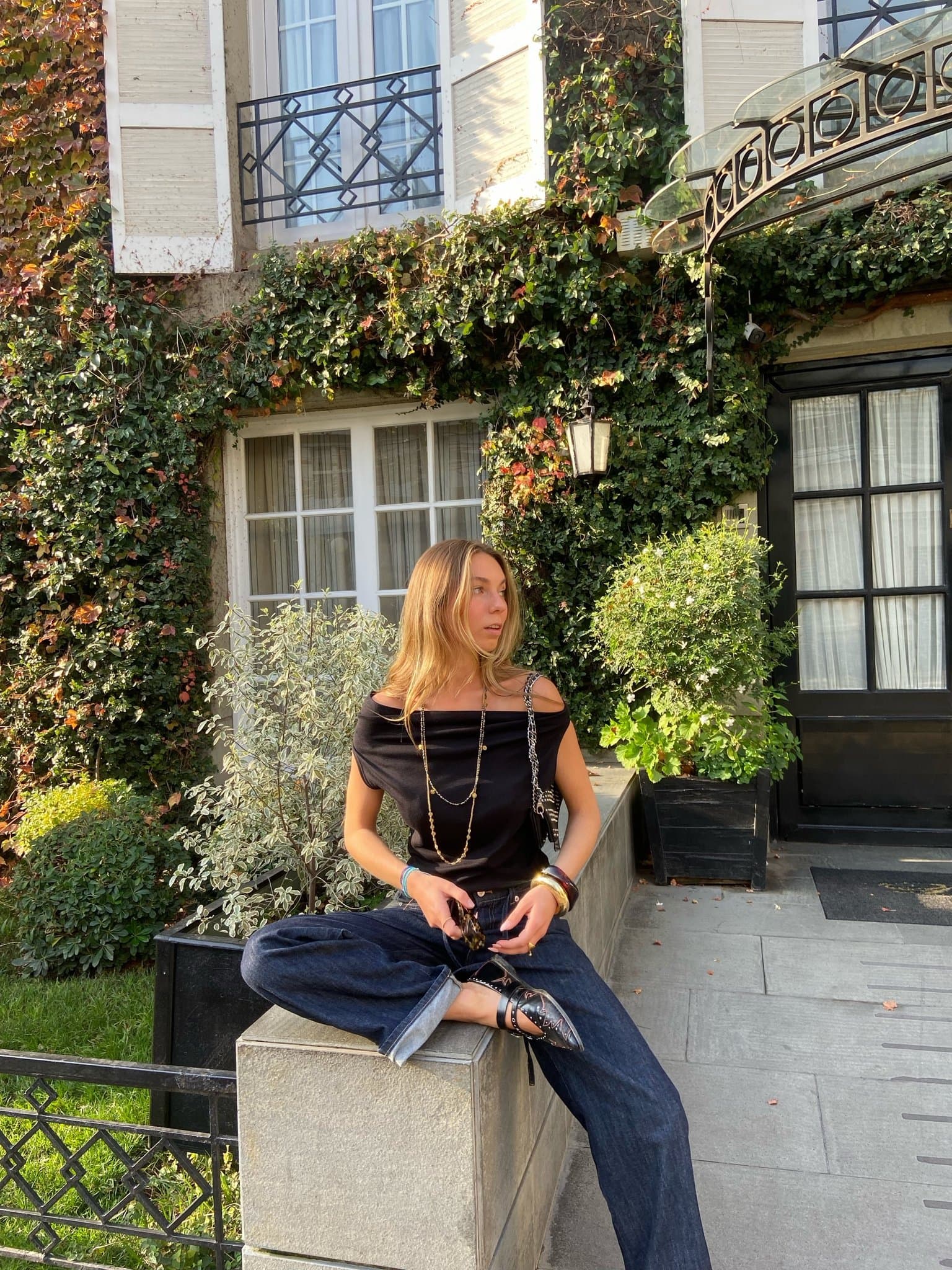 A black off-shoulder top is styled with dark wide-leg cuffed jeans, pointed studded strap flats, layered gold jewelry, and a small black chain-strap bag.