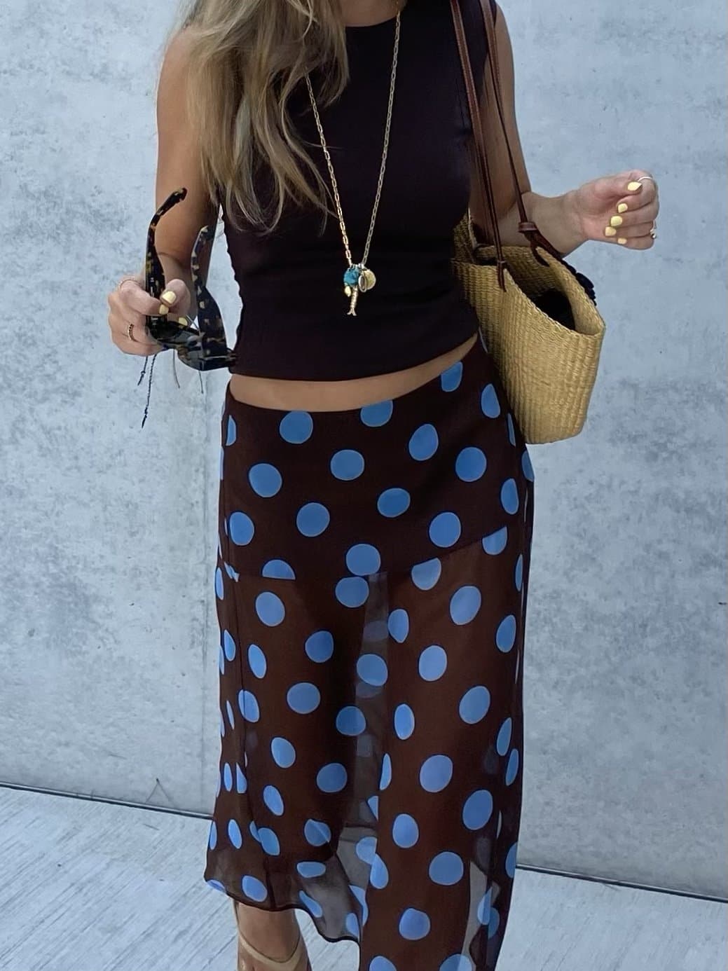 A dark sleeveless crop top is styled with a sheer brown blue-polka-dot midi skirt, layered necklaces, sunglasses, and a woven shoulder bag.
