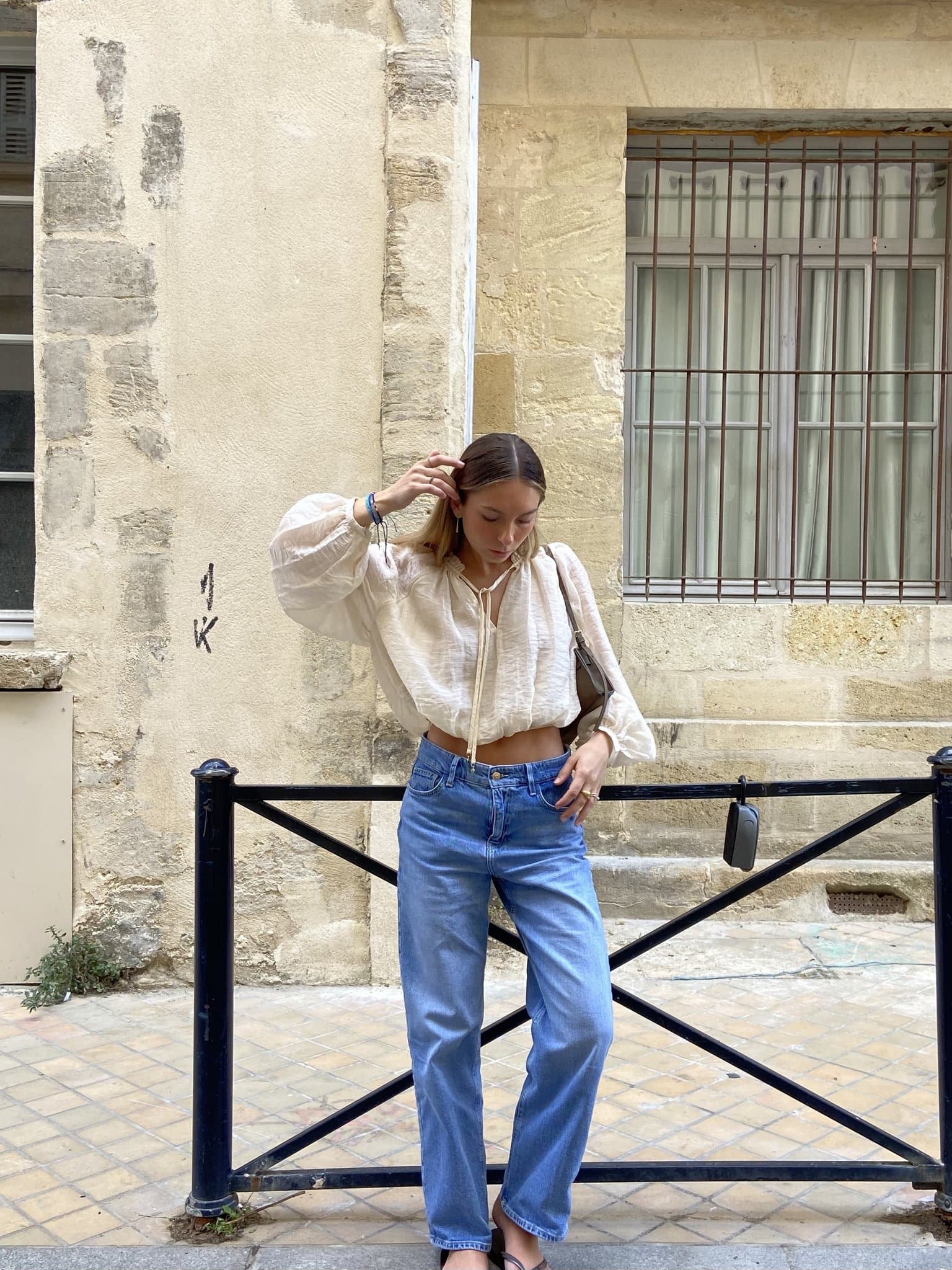 A cropped white voluminous blouse is styled with light blue straight-leg jeans, black flat sandals, and a taupe shoulder bag.