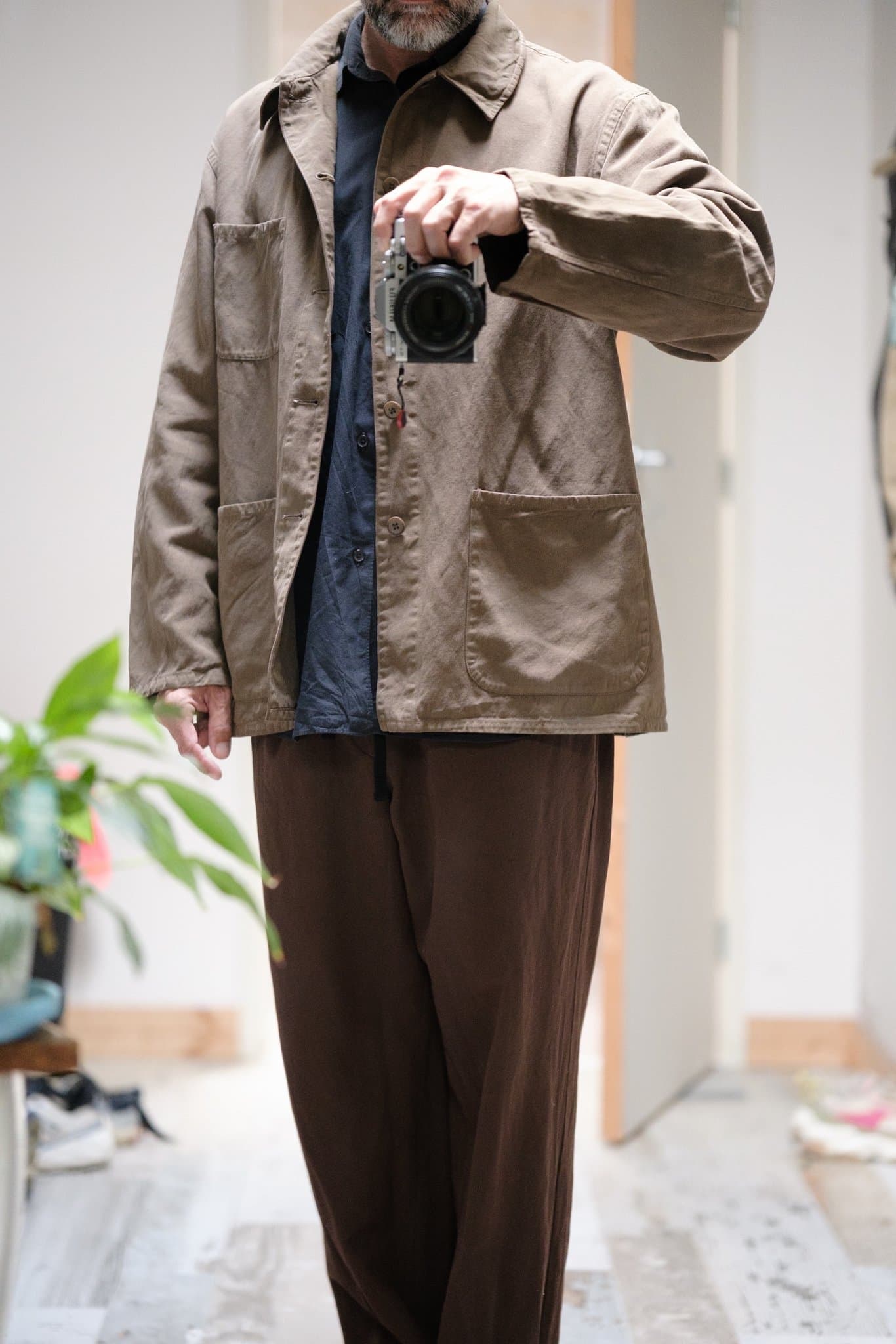 A taupe chore jacket is layered over a dark navy shirt with loose brown trousers.