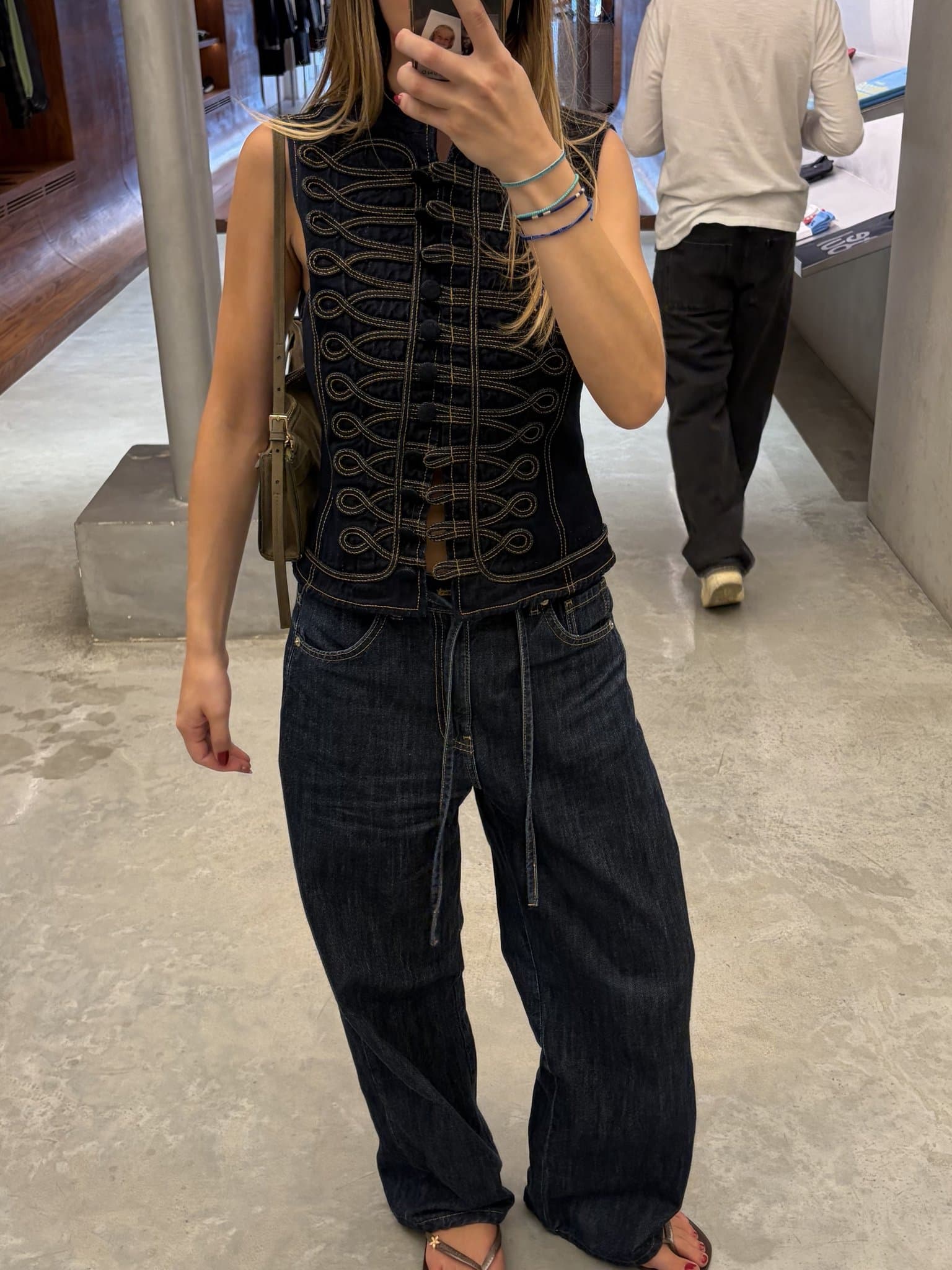 A dark denim sleeveless military-style vest is paired with wide dark denim jeans, slim sandals, a shoulder bag, and stacked cord bracelets.