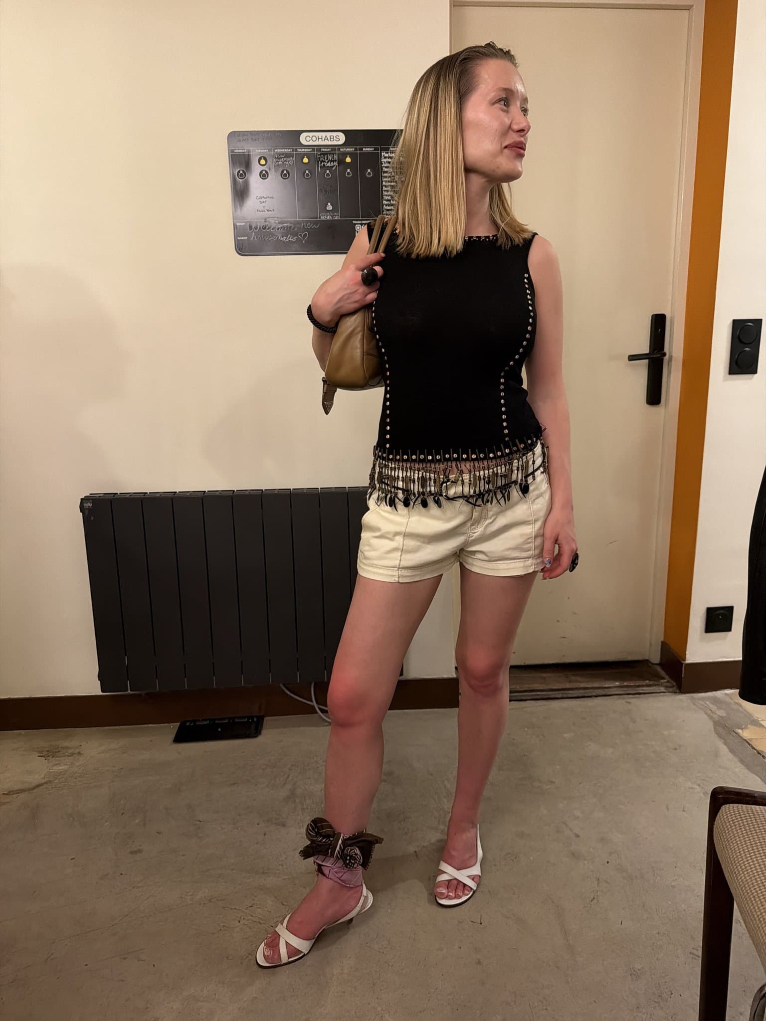 A black embellished sleeveless top with beaded fringe is styled with cream shorts, a tan shoulder bag, ankle-tied scarf detail, and white heeled sandals.