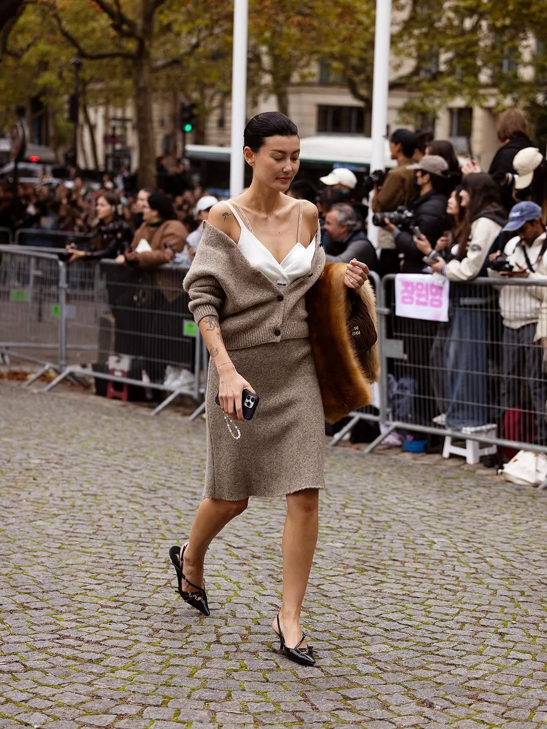 A taupe knit cardigan and matching knee-length skirt are worn off the shoulder over a white bralette, with black pointed slingbacks and a fur stole carried at the side.