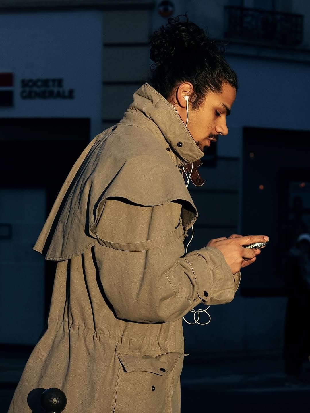 A tan oversized utility trench coat with a high collar, cape-like shoulder flap, and gathered waist is worn with white wired earbuds.