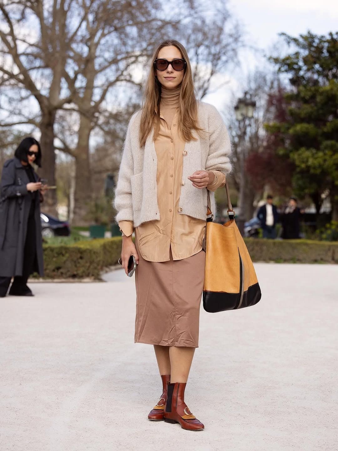 A neutral layered look with a cream fuzzy cardigan, tan shirt, brown midi skirt, color-block tote, and polished brown ankle boots.
