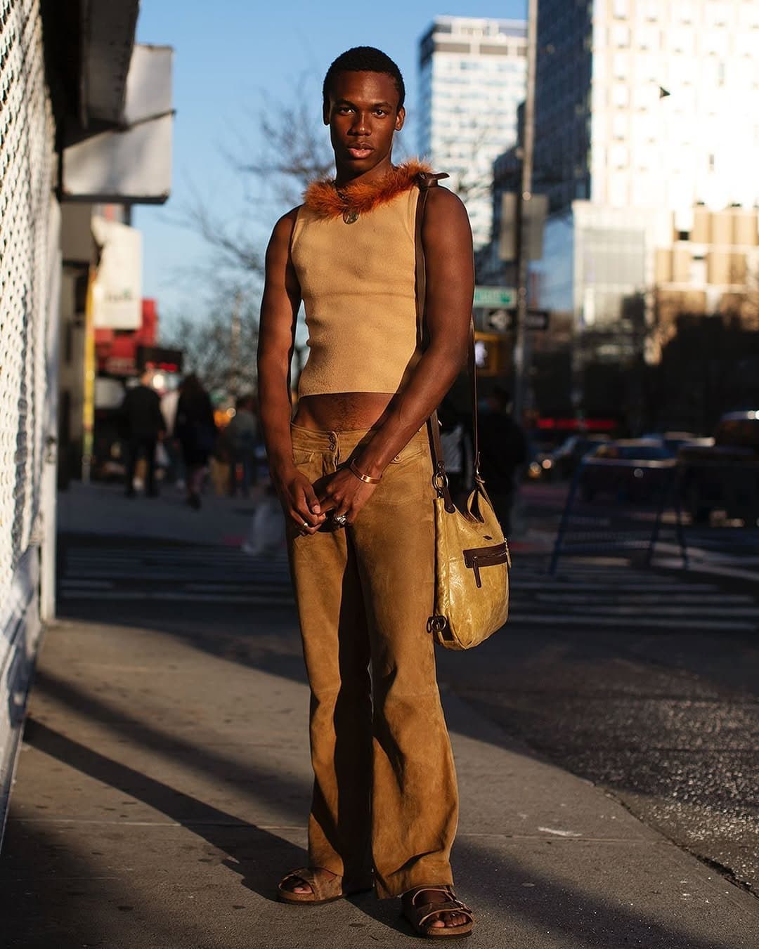 A tonal tan street look pairs a cropped sleeveless knit top with fur trim, low-rise flared pants, brown sandals, and a slouchy shoulder bag.
