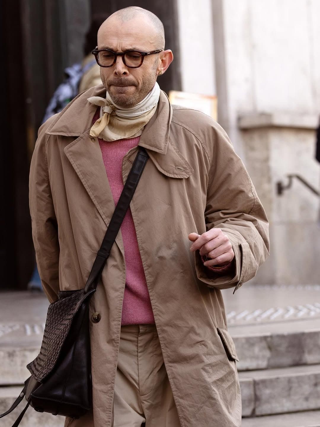 A beige oversized trench coat is layered over a pink knit with beige trousers, a tied neck scarf, glasses, and a black crossbody bag.