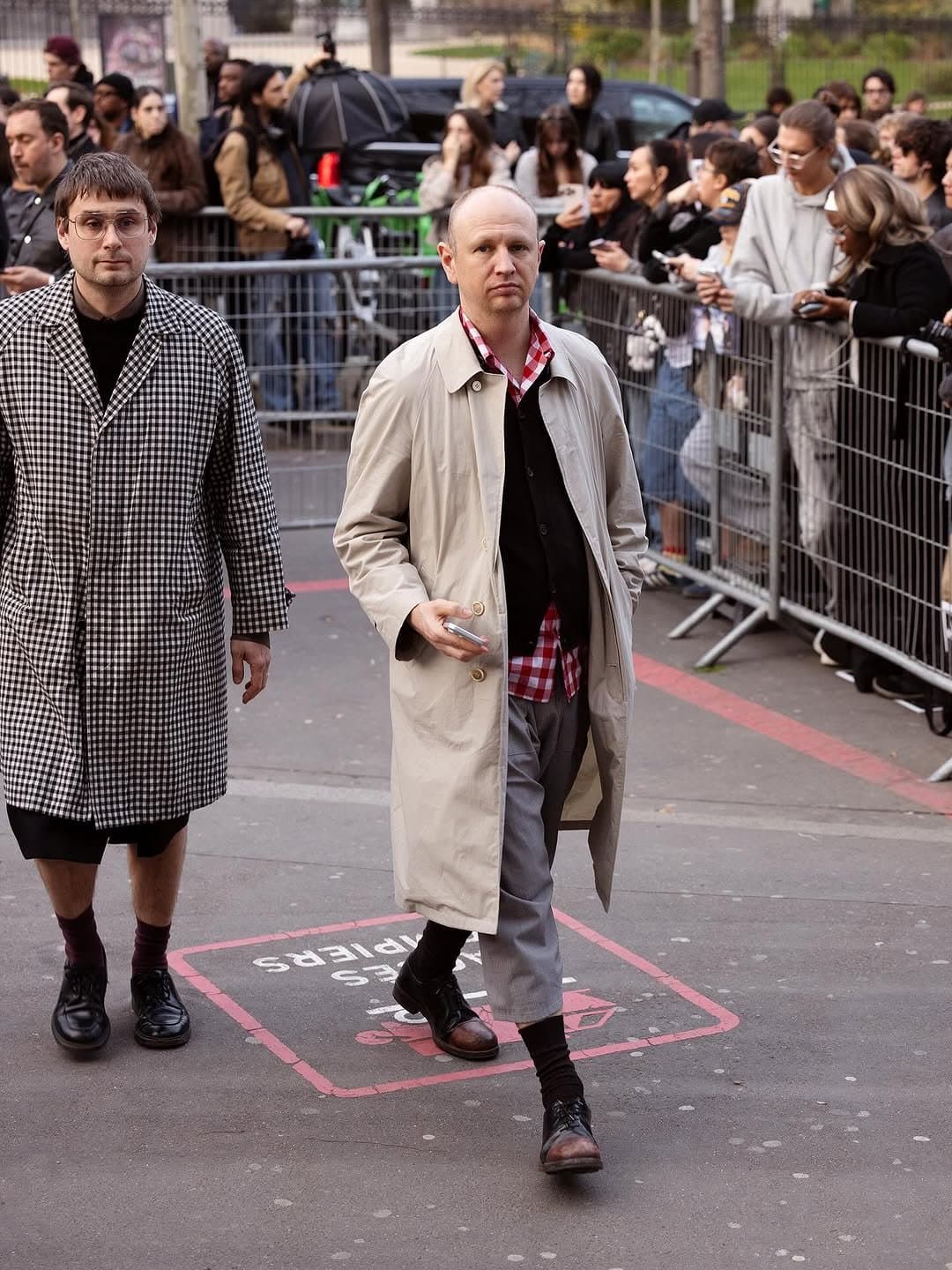 A beige trench coat is layered over a black cardigan, red gingham shirt, grey trousers, black socks, and worn leather boots.