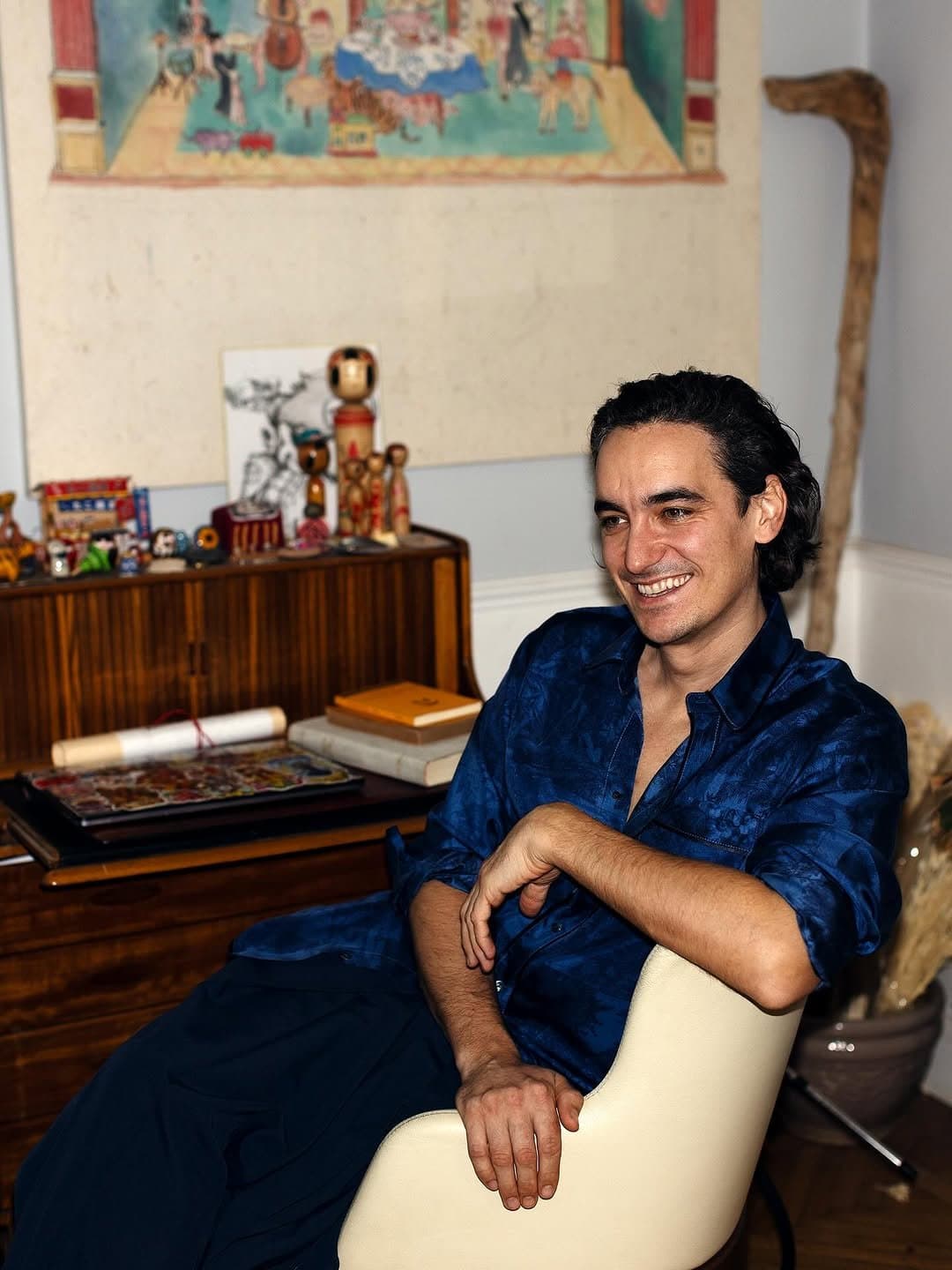A man sits in a deep blue patterned button-up shirt with dark blue trousers.