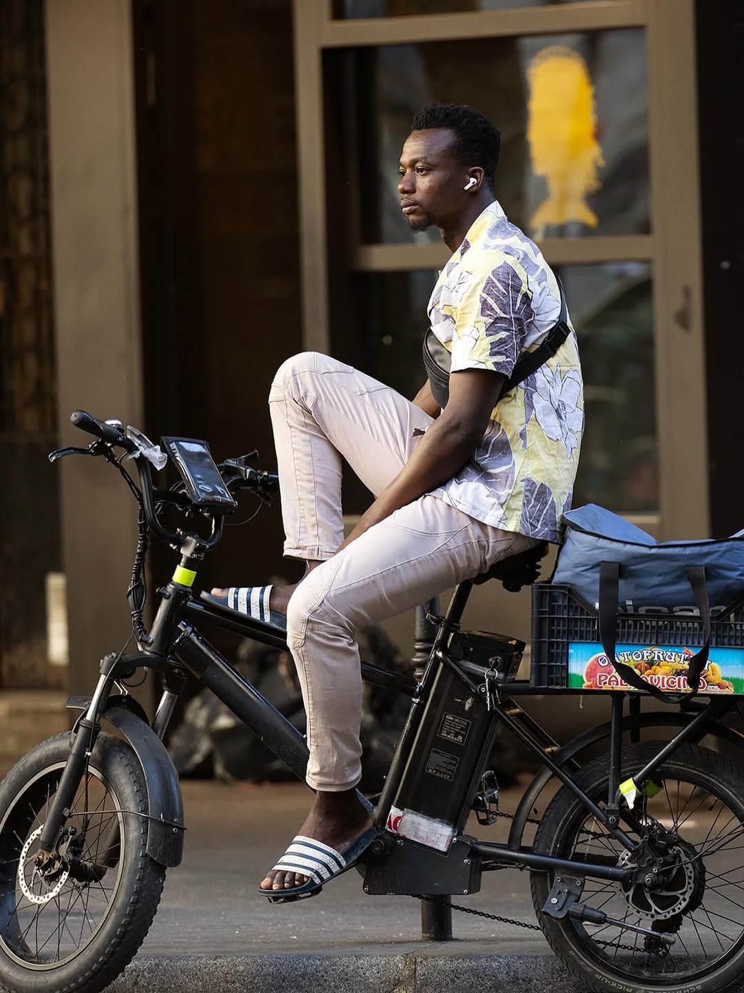 A floral short-sleeve shirt is worn with pale slim trousers, striped slide sandals, and a black crossbody chest bag.