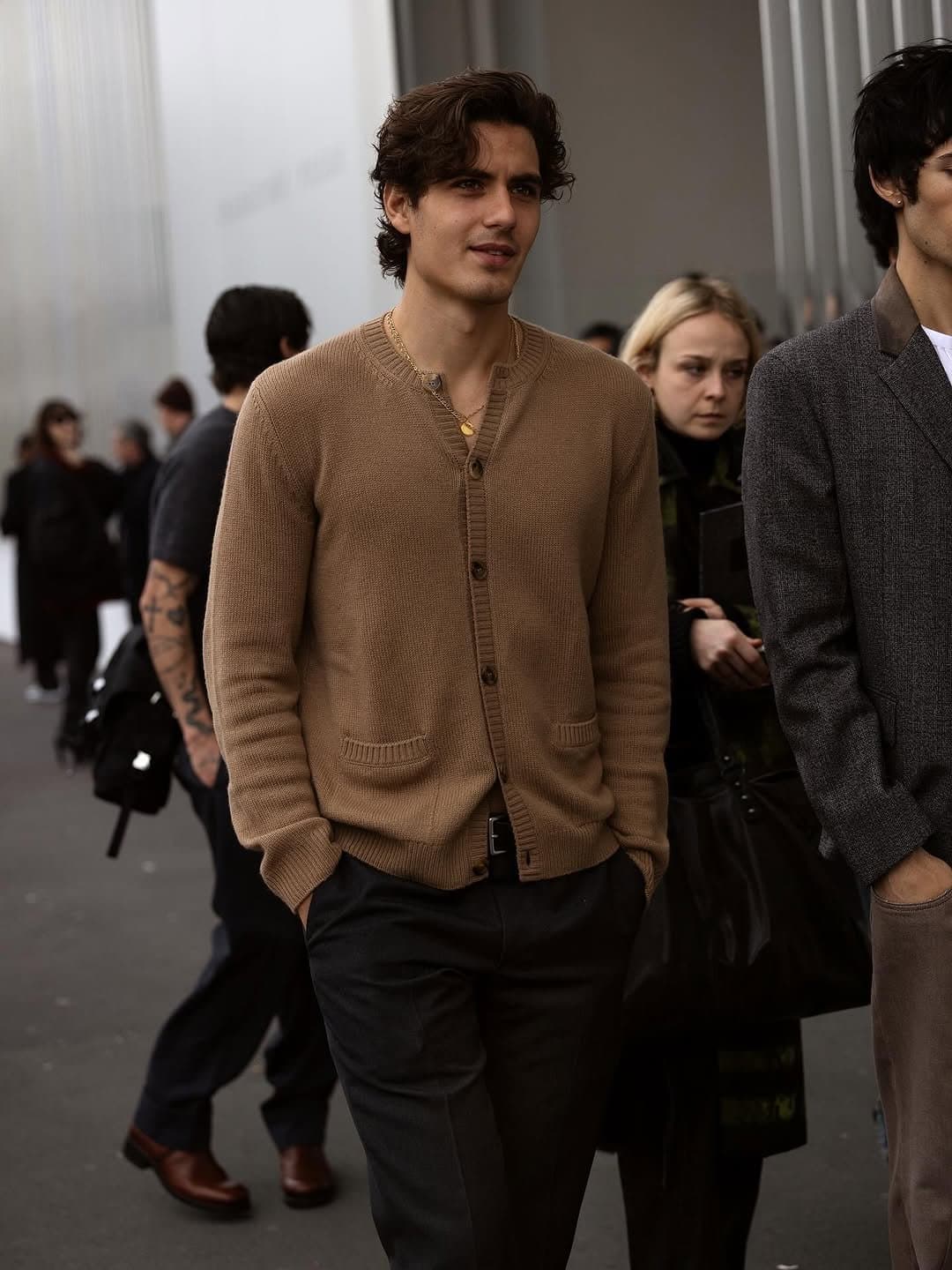 A tan knit cardigan is worn buttoned over dark tailored trousers with a black belt and layered gold necklaces.