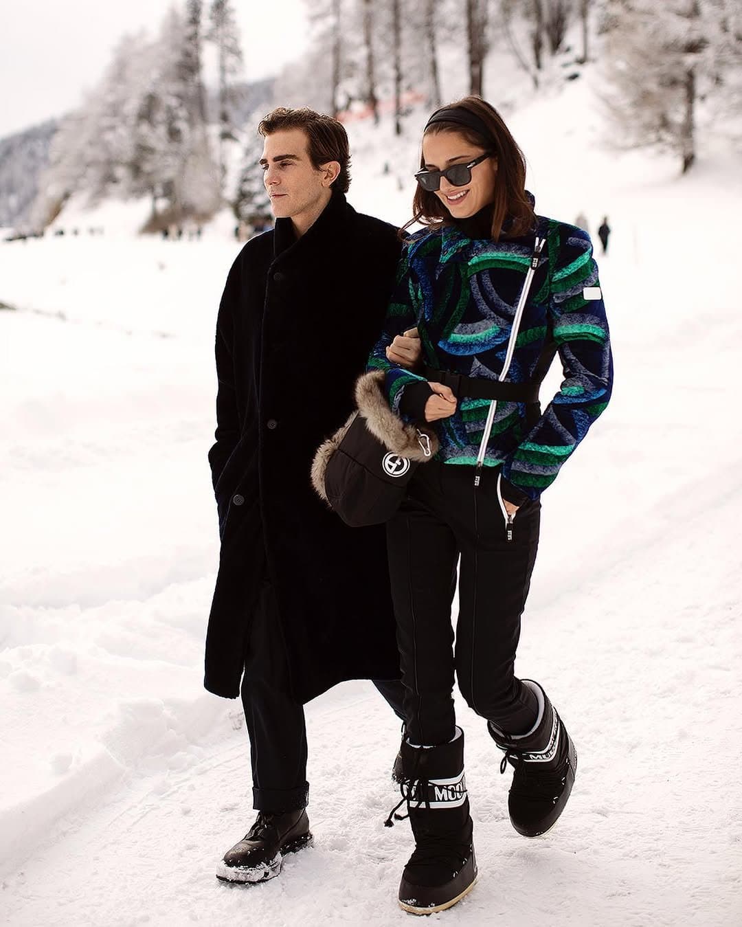 A winter ski look with a bold blue-green patterned jacket, black snow pants, black moon boots, sunglasses, and a fur-trimmed black bag.