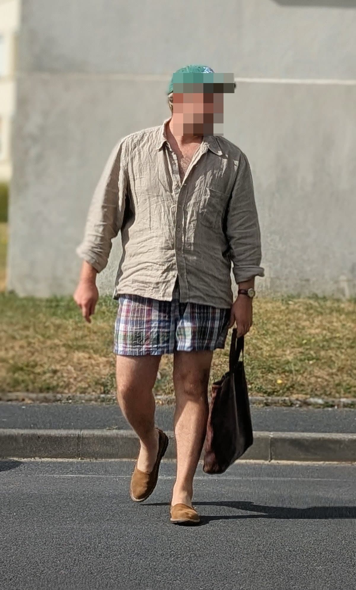 A relaxed beige crinkled button-up shirt is worn with multicolor plaid shorts, tan slip-on shoes, a wristwatch, and a dark tote bag.