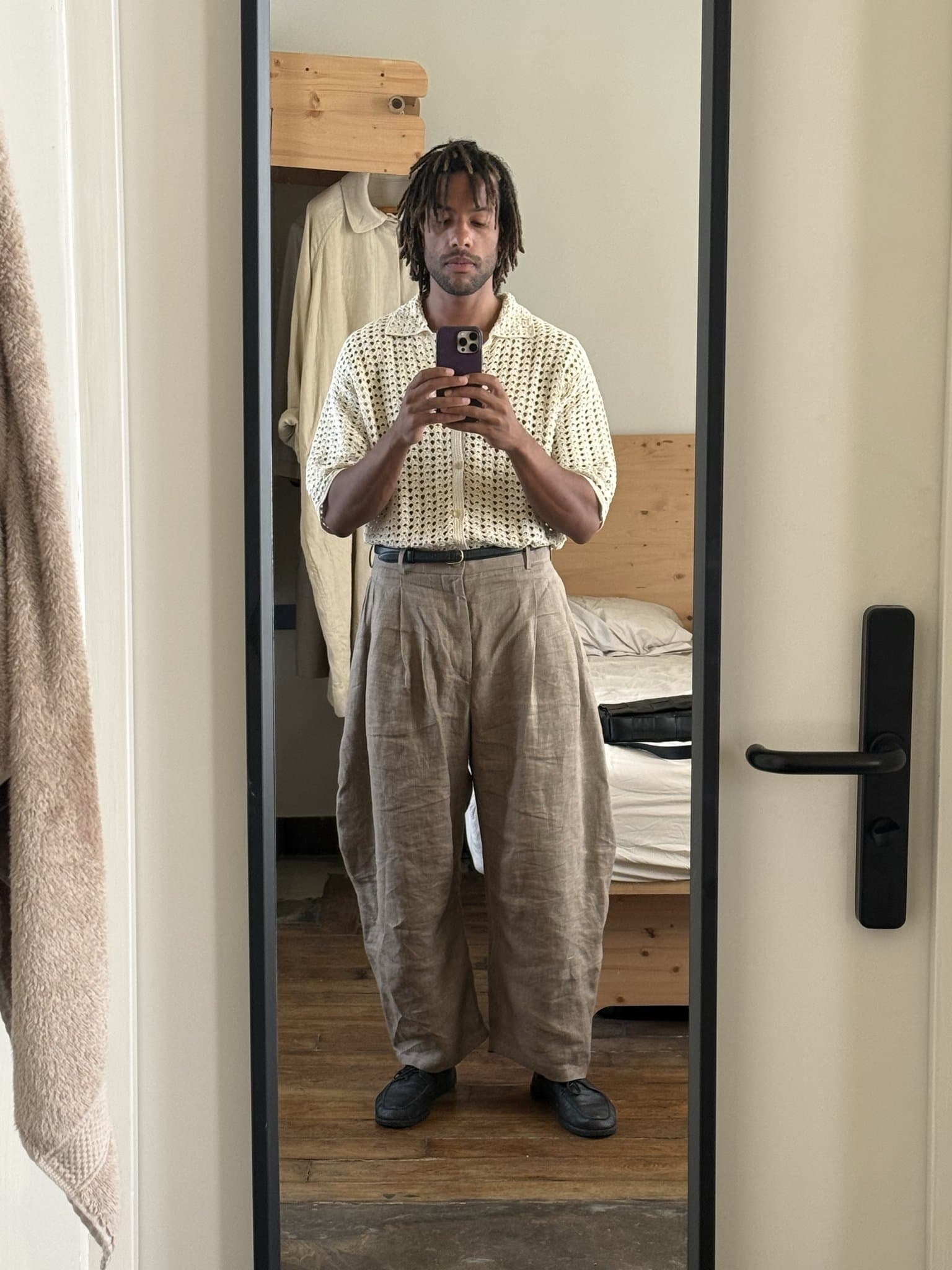 Cream open-knit collared shirt tucked into voluminous beige pleated trousers with a black belt and black shoes.