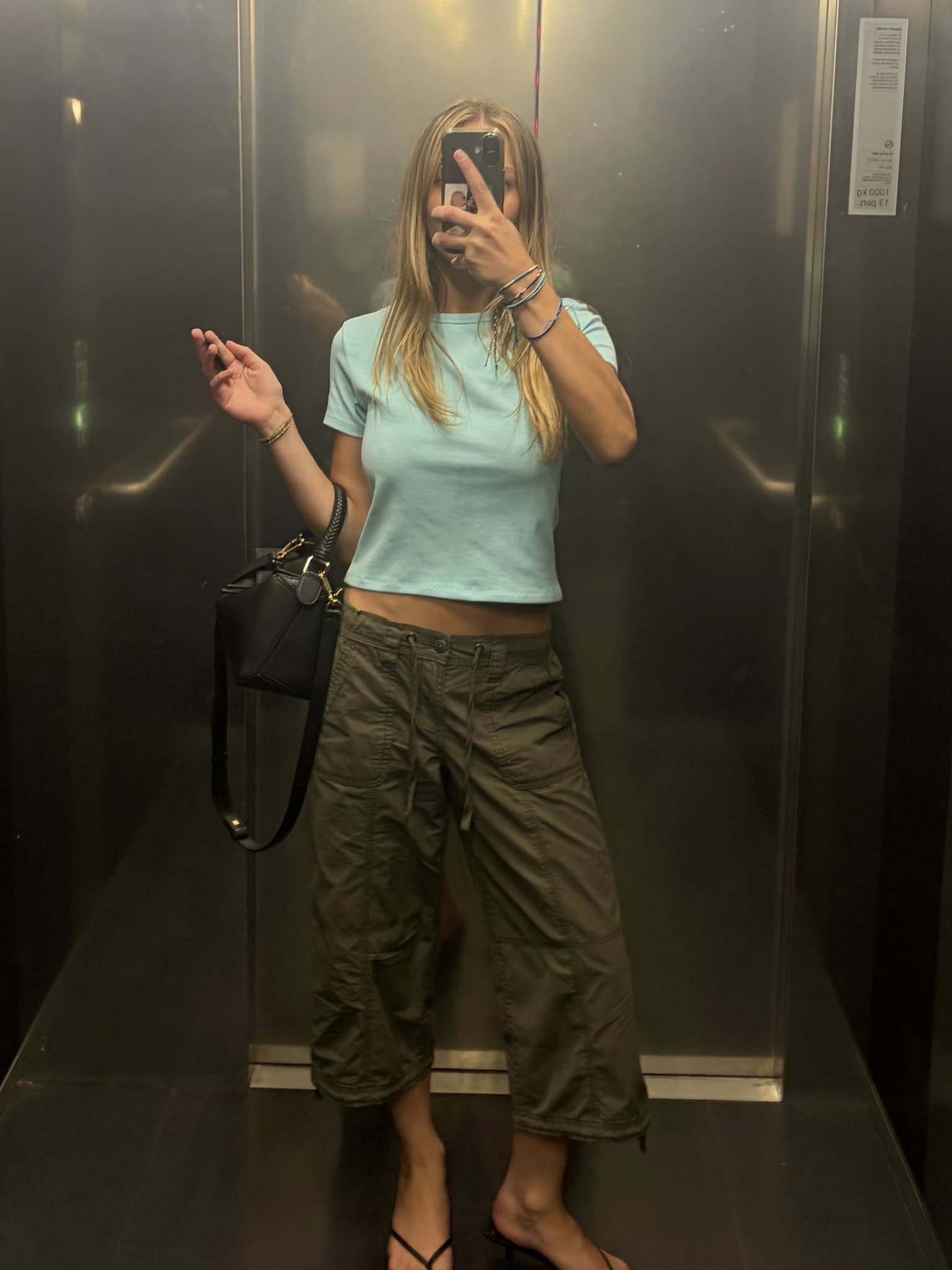 A pale aqua cropped tee is styled with oversized olive cargo trousers, black strappy heels, a black shoulder bag, and stacked bracelets.