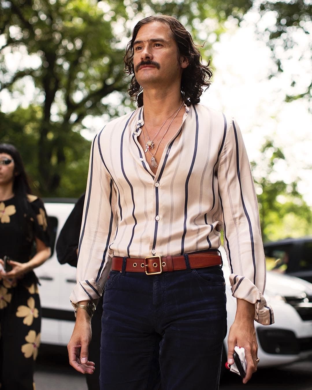 A cream vertical-striped open-neck shirt is tucked into dark navy trousers with a reddish brown leather belt and layered pendant necklaces.