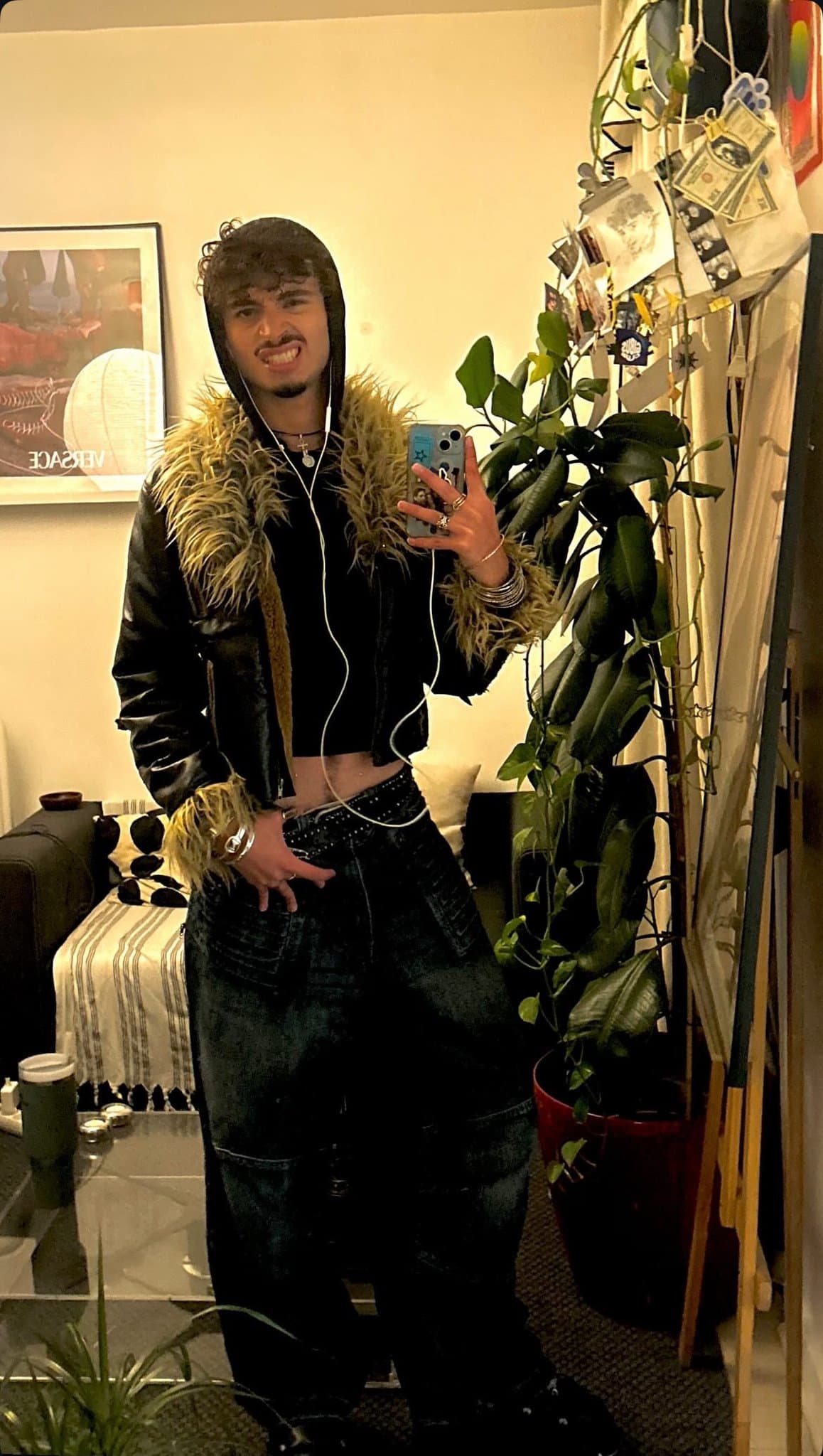 A black hooded faux-fur-trim jacket is worn open over a cropped black top with dark relaxed denim, a studded belt, and silver jewelry.