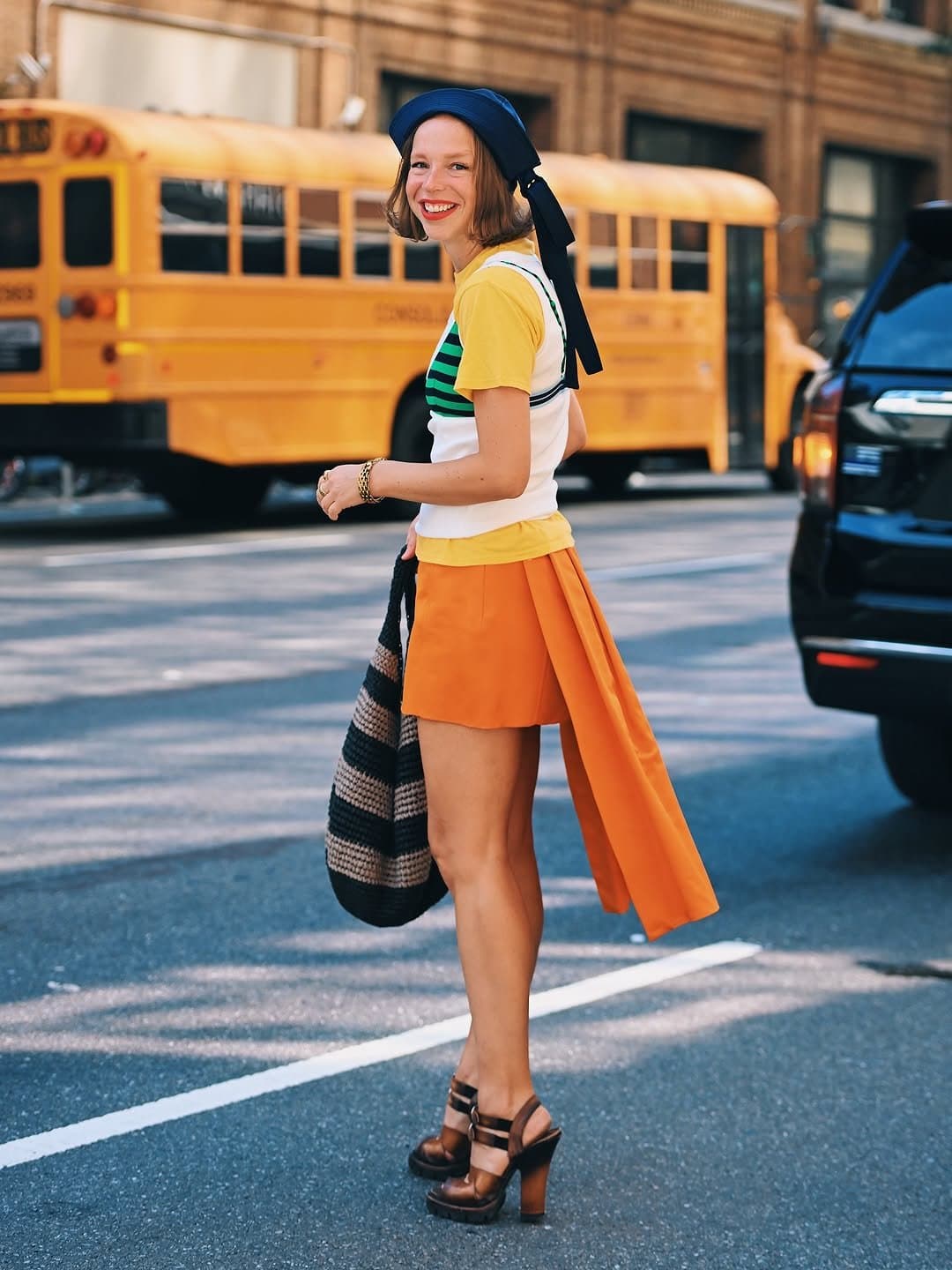 A colorful layered street-style look with a yellow tee, striped knit vest, orange mini skirt, navy headscarf, striped bag, and chunky brown platform sandals.