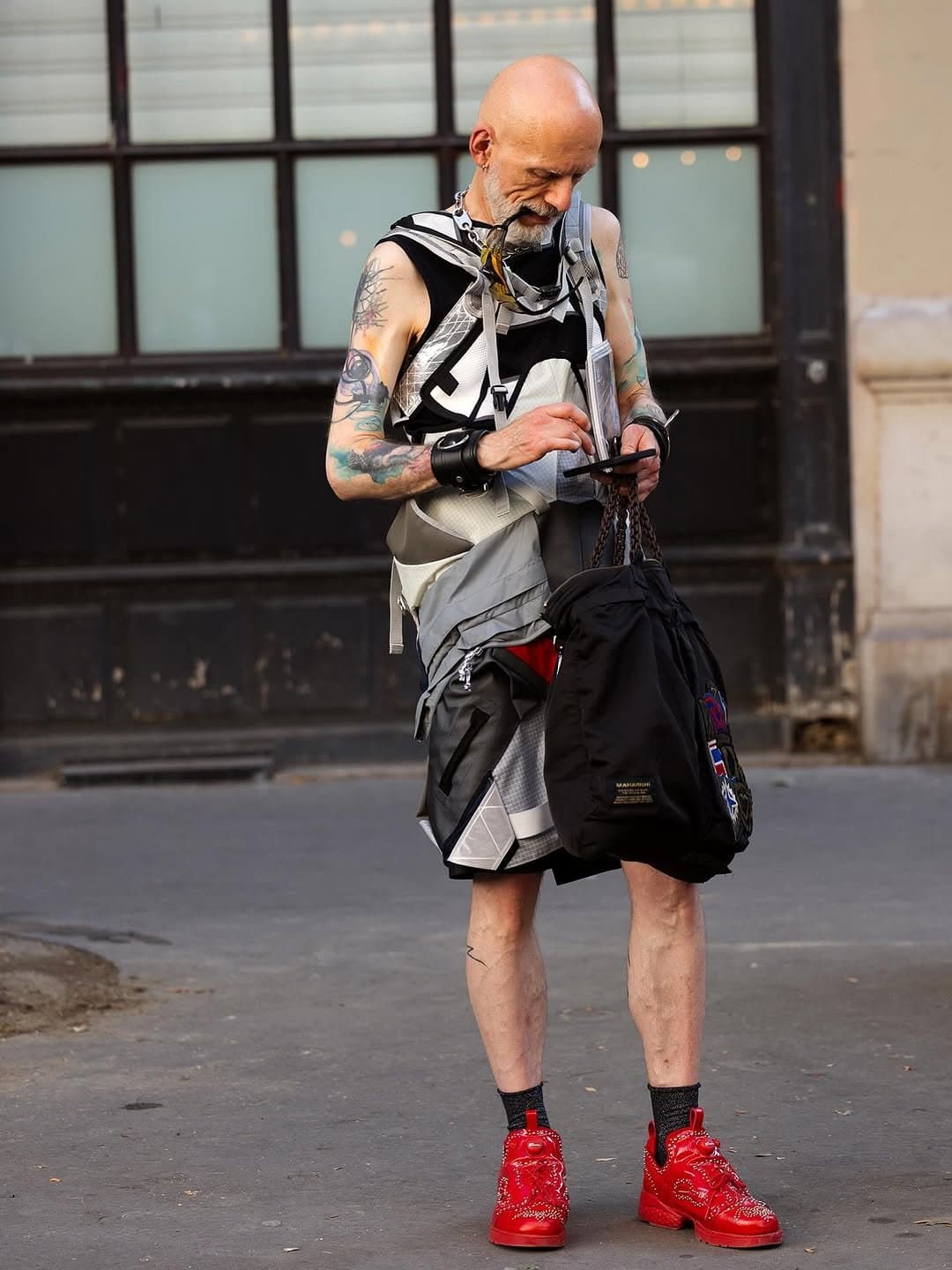 A layered technical black, grey, and white outfit is styled with a neck scarf, black utility bag, wrist cuffs, and glossy red high-top sneakers.