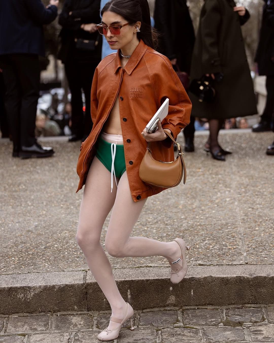 A cognac leather jacket is styled with green high-cut briefs, pale pink tights, Mary Jane heels, tinted sunglasses, and a tan shoulder bag.