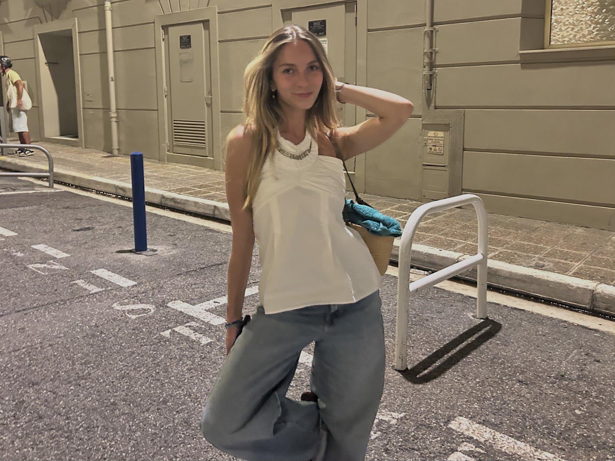 A white sleeveless halter-style top is styled with relaxed light blue-grey wide-leg trousers, metallic jewelry, and a woven shoulder bag.