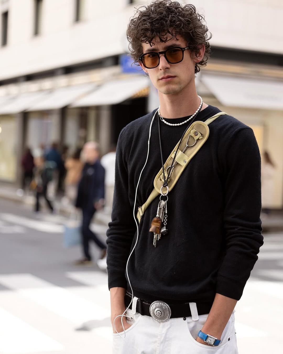 Black crewneck knitwear is tucked into white trousers with a western buckle, pearl-like necklace, sunglasses, and a tan utility crossbody strap.