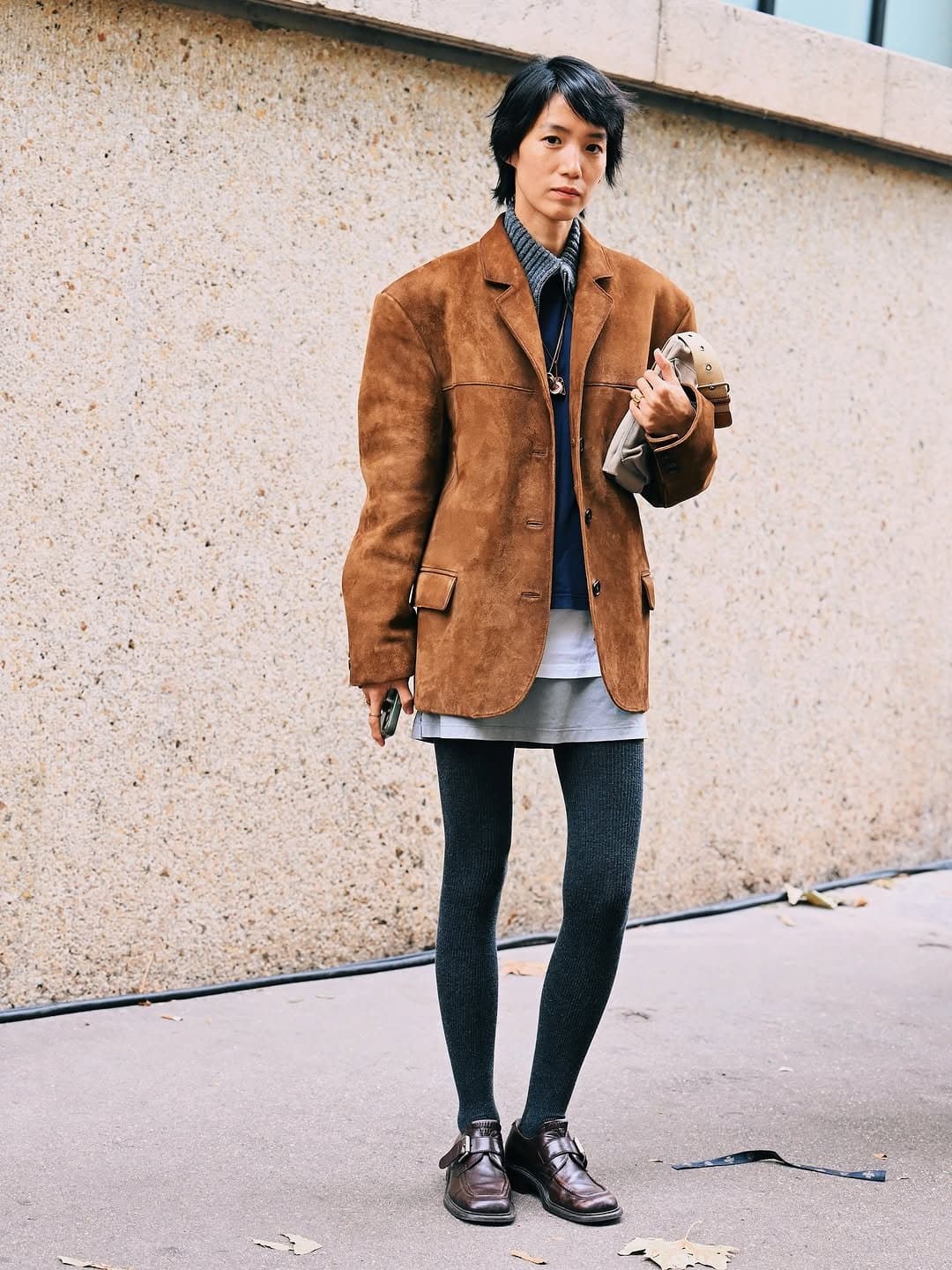 A brown suede blazer is layered over grey and navy pieces with a short grey skirt, dark ribbed tights, and polished dark loafers.