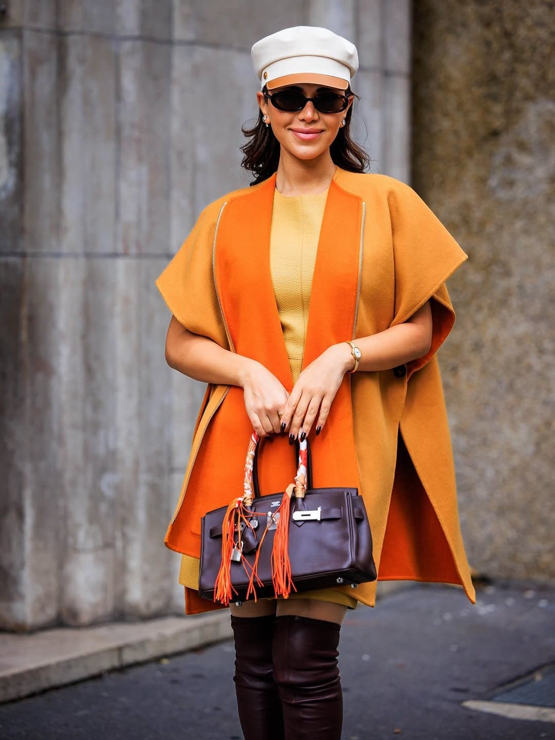 Camel and orange cape styling with a mustard mini dress, cream cap, dark sunglasses, structured brown handbag, and tall brown boots.