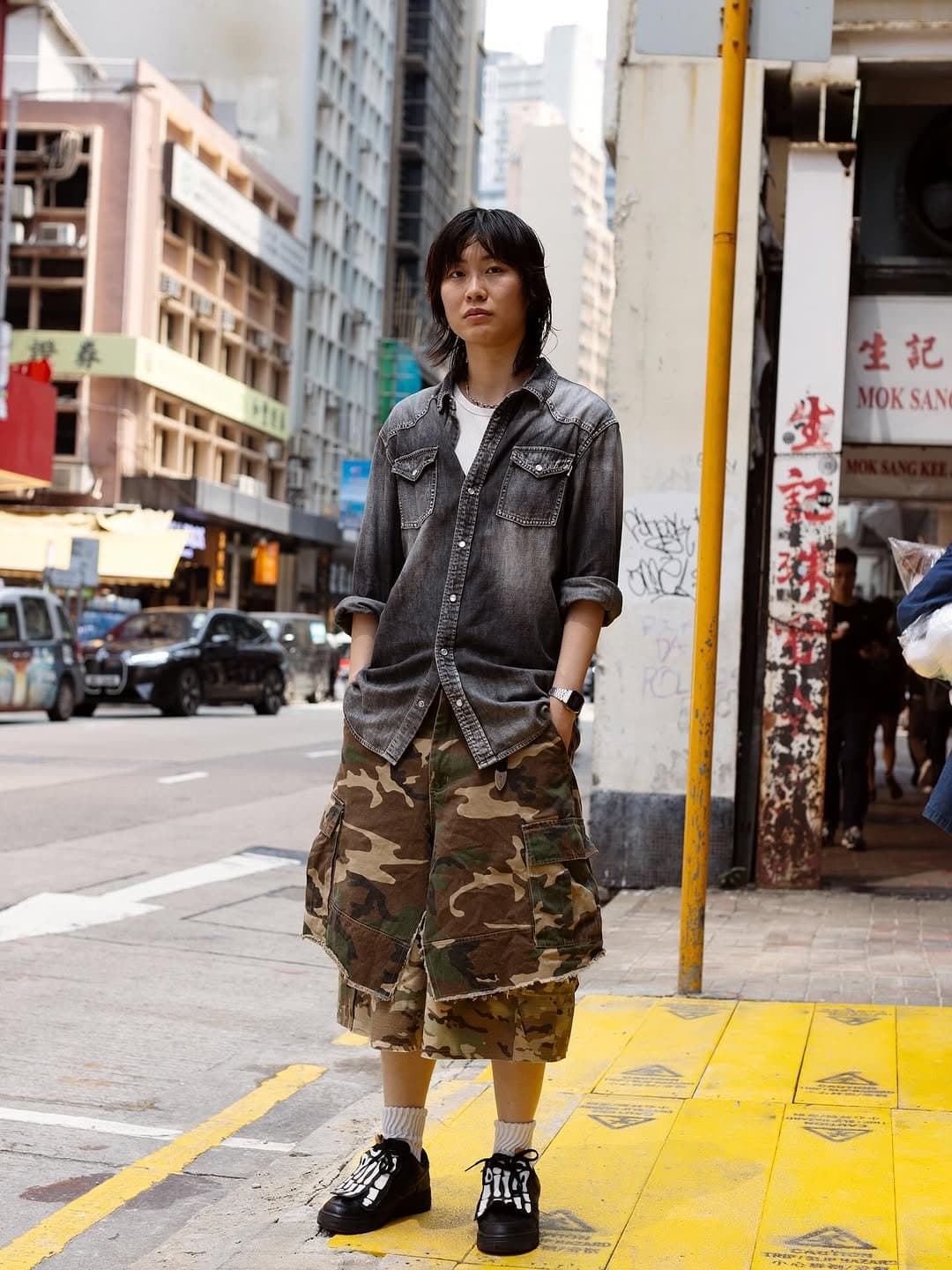 A relaxed grey denim shirt is styled with long camouflage cargo shorts, white socks, and black graphic sneakers.