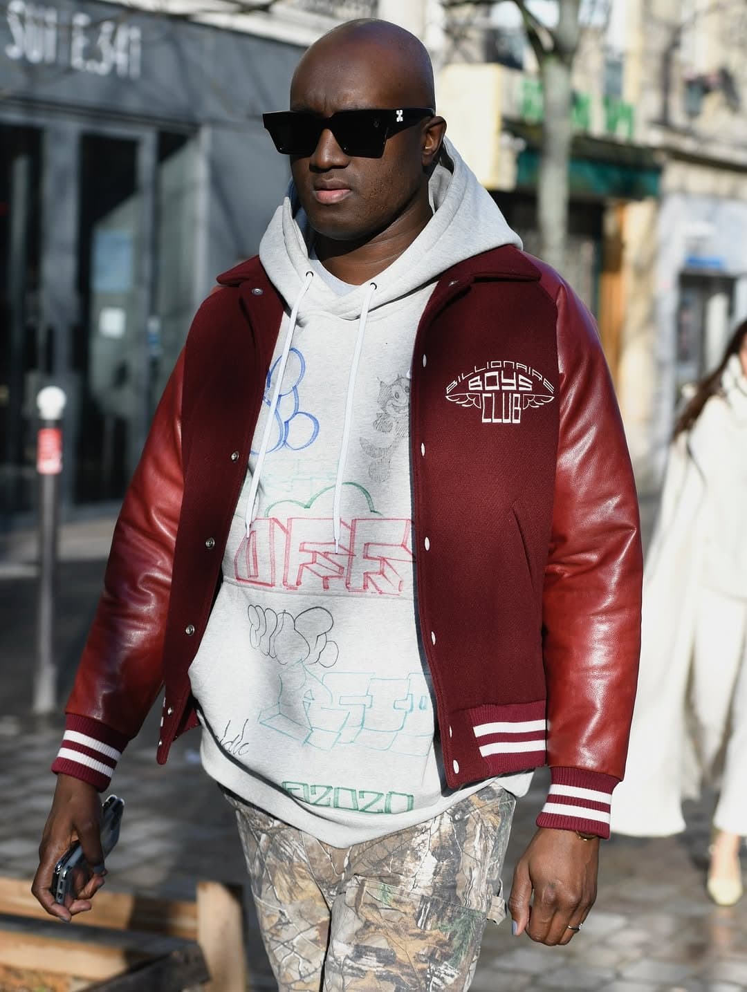 A burgundy varsity jacket is layered over a grey graphic hoodie with camouflage cargo-style pants and black rectangular sunglasses.