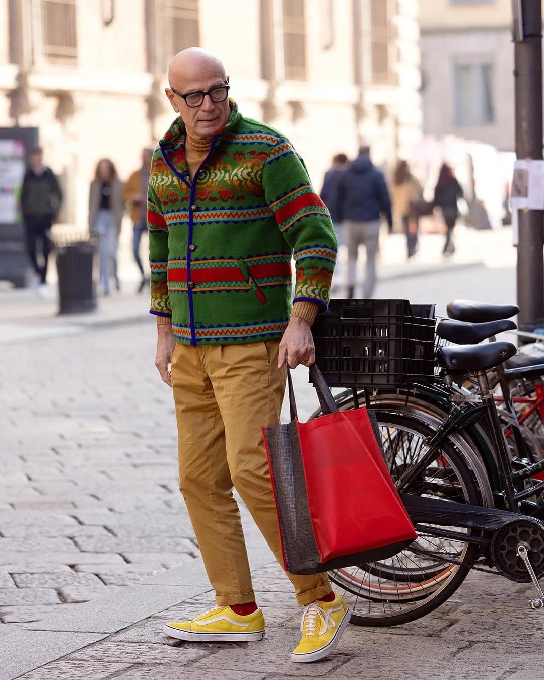 A colorful patterned green jacket is worn with a mustard turtleneck, tan chinos, yellow sneakers, red socks, and a red tote.