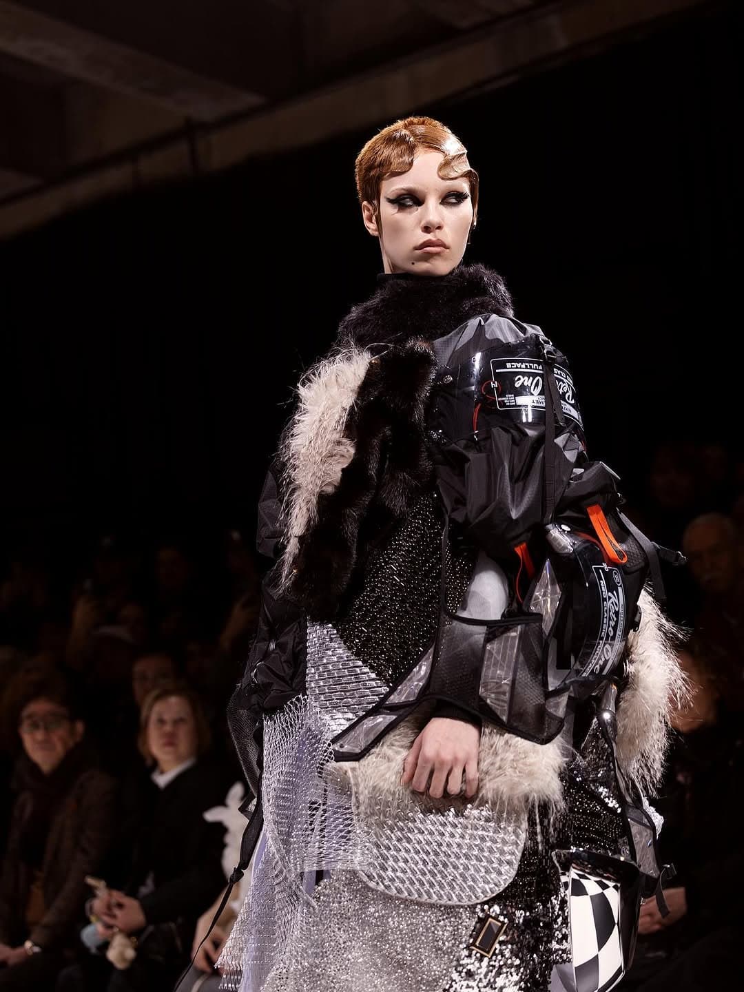 A layered runway look combines black technical panels, shaggy fur textures, sequined black surfaces, metallic silver mesh, and graphic checkerboard accents.
