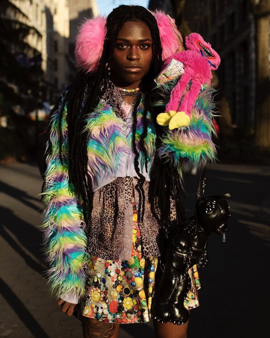 A maximal multicolor faux-fur look with layered prints, pink fluffy earmuffs, a bright mini skirt, and glossy black accessories.