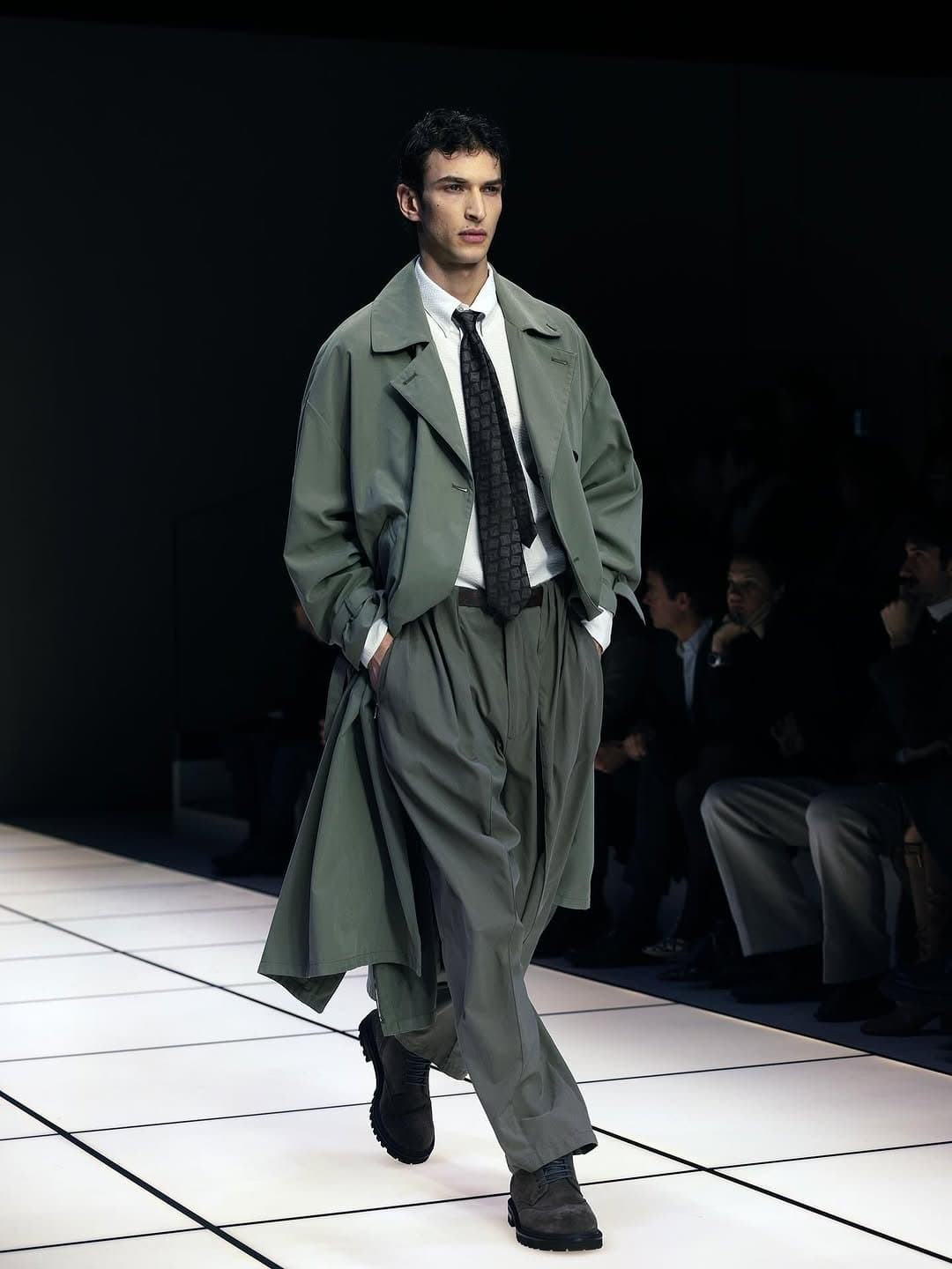 An oversized sage trench-style coat is layered over a white shirt, dark tie, wide pleated trousers, and black lug-sole boots.
