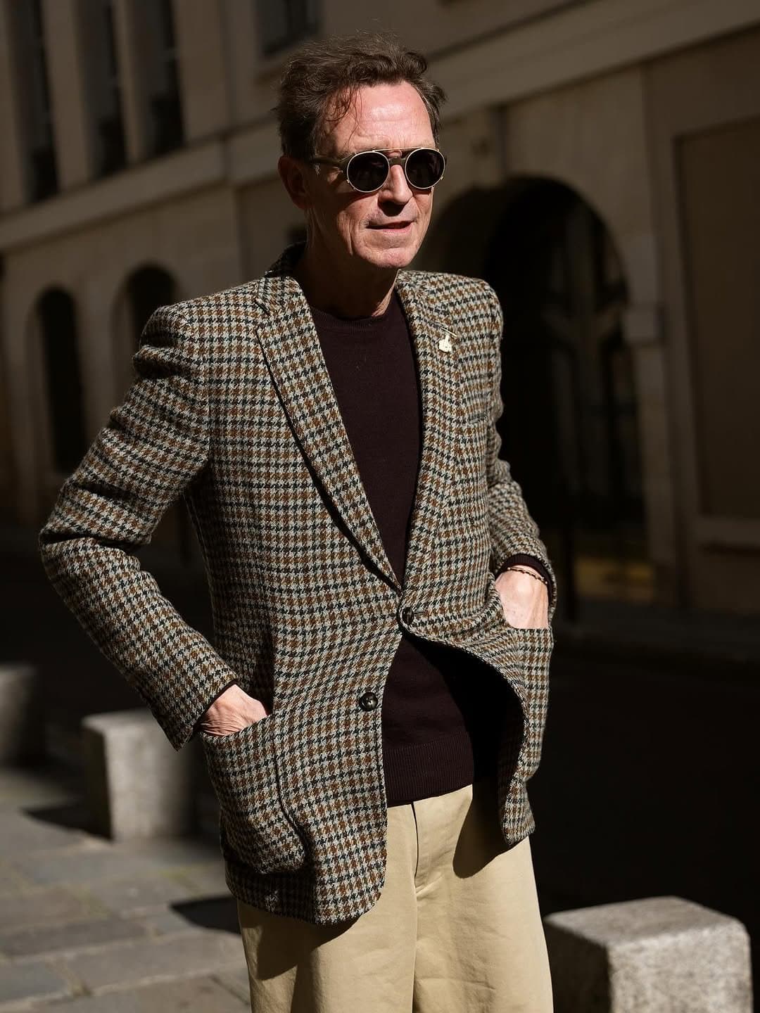 A houndstooth tweed blazer is worn over a dark brown crew neck knit with beige relaxed trousers and round sunglasses.