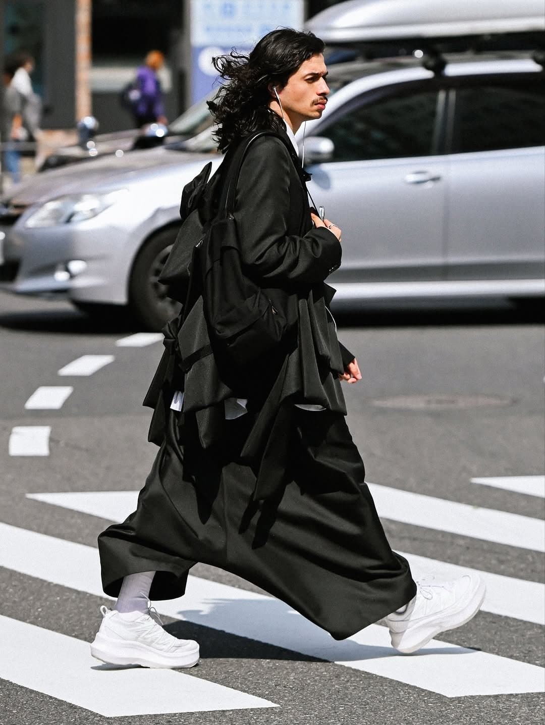 A black oversized layered outfit with wide draped trousers, white shirt accents, white chunky sneakers, and black bags.