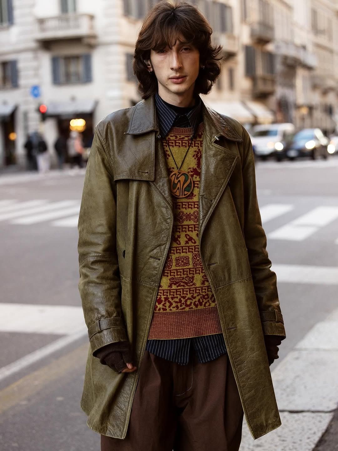 An olive leather coat is layered over a patterned knit vest, striped shirt, loose brown trousers, knit gloves, and a bold pendant necklace.