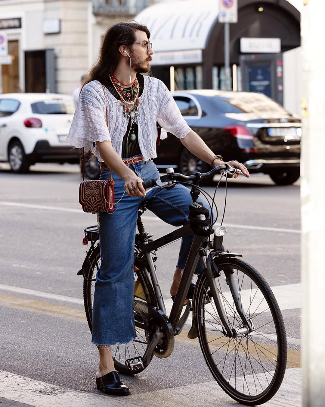 A white cropped lace top over black knit, wide cropped blue jeans, black mules, a red patterned bag, and layered jewelry.