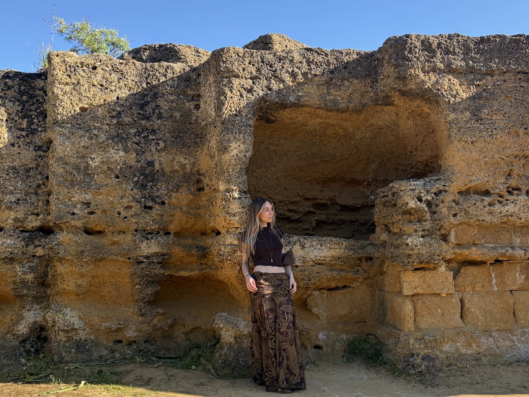A fitted dark cropped tee is paired with flowing patterned wide-leg trousers, a small shoulder bag, and a long pendant necklace.