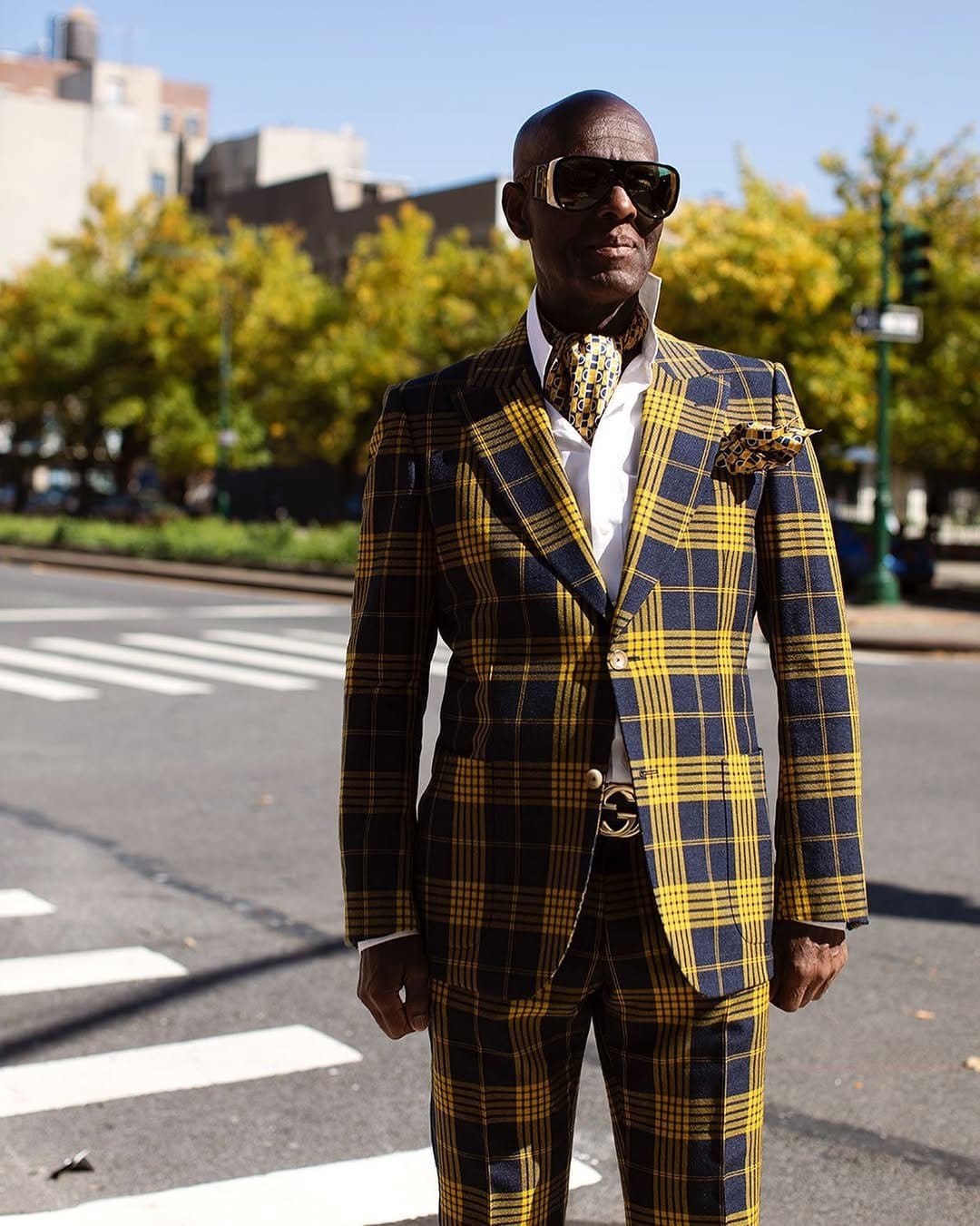 A tailored navy-and-yellow plaid suit is styled with a white shirt, matching printed scarf and pocket square, oversized sunglasses, and a gold-buckled belt.