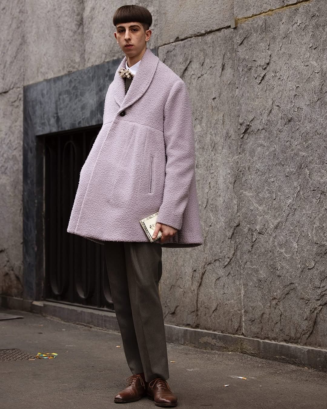 A pale pink textured oversized coat is styled with tailored dark trousers, brown leather lace-up shoes, and a cream neck bow.