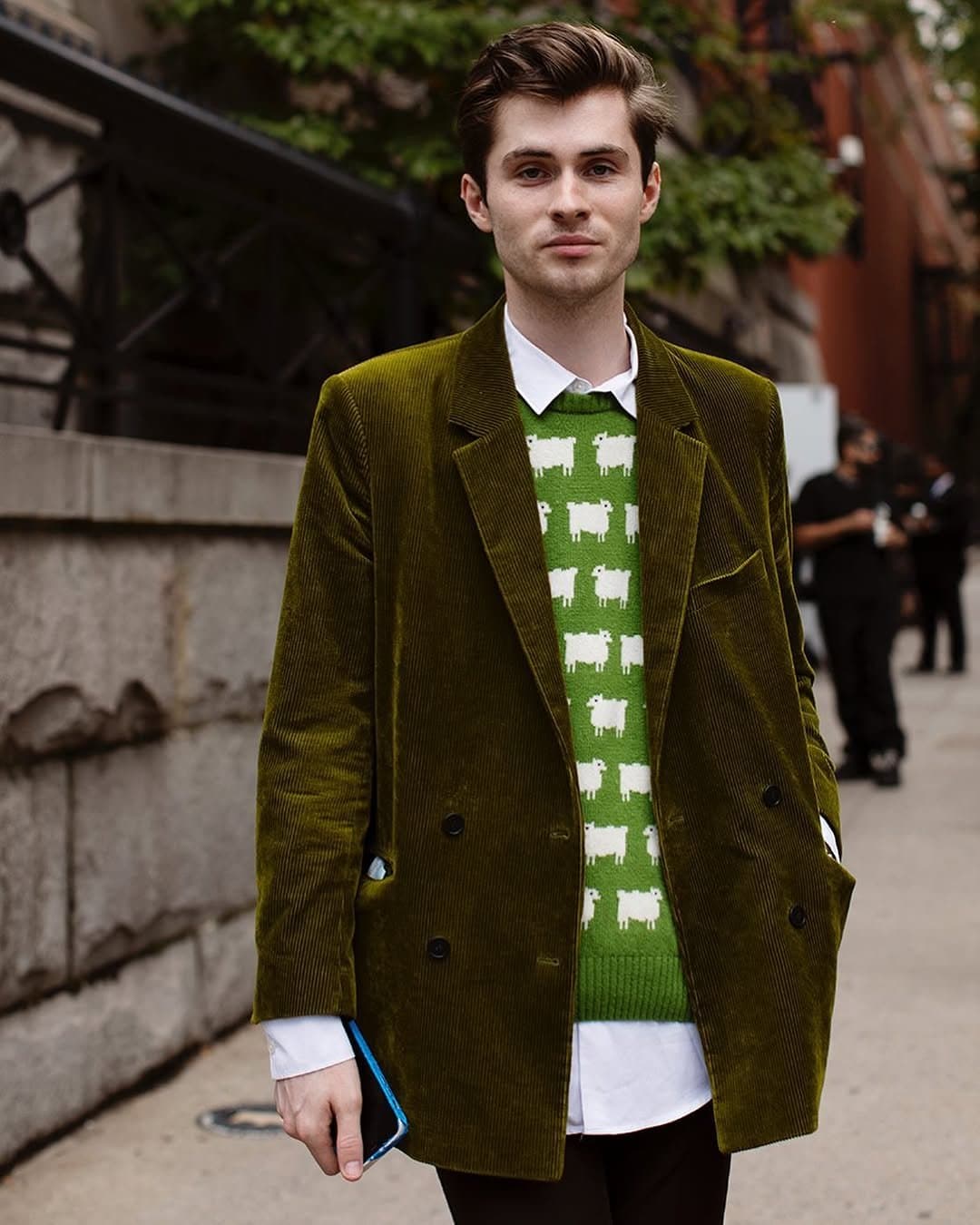 A green corduroy blazer is layered over a white shirt and a bright green sheep-pattern knit vest with dark trousers.