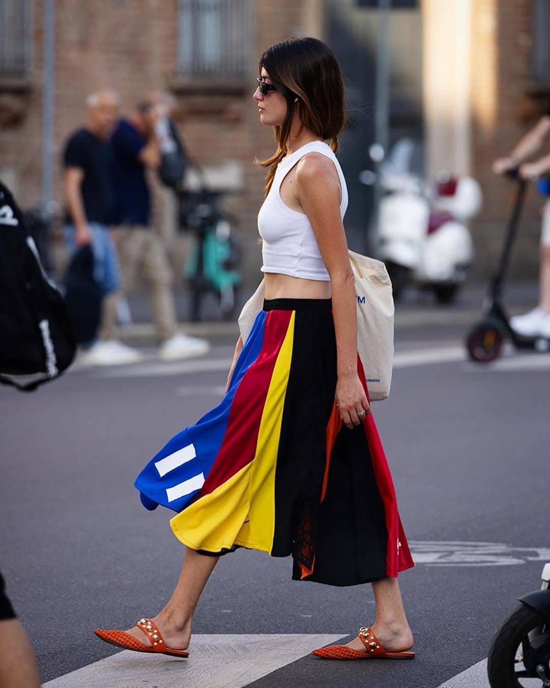 A white cropped sleeveless top is styled with a bold multicolor midi skirt, orange embellished slides, black sunglasses, and a cream canvas tote.