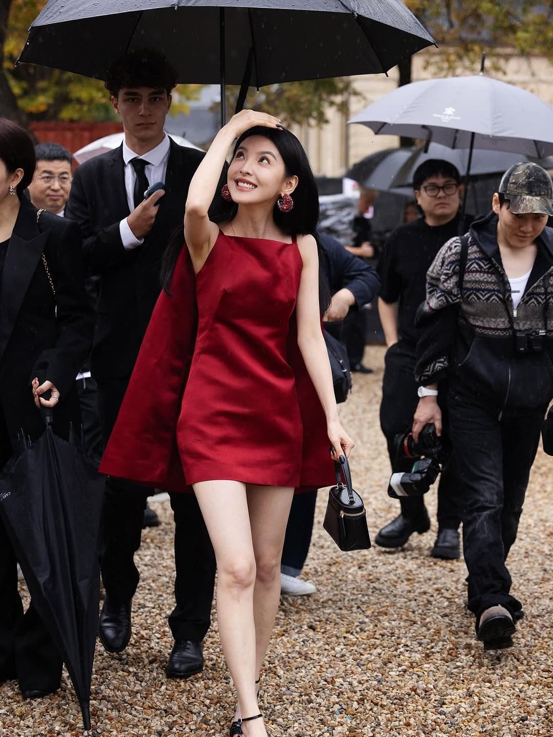 A deep red mini dress with a dramatic one-sided cape panel is styled with red statement earrings, a black mini bag, and black ankle-strap heels.